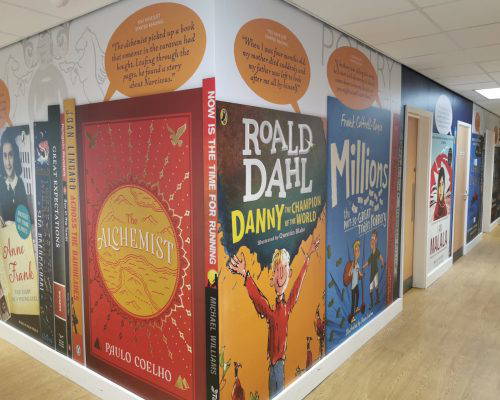 School library hallway with book cover murals: Anne Frank, The Alchemist, Danny the Champion of the World, and Millions.